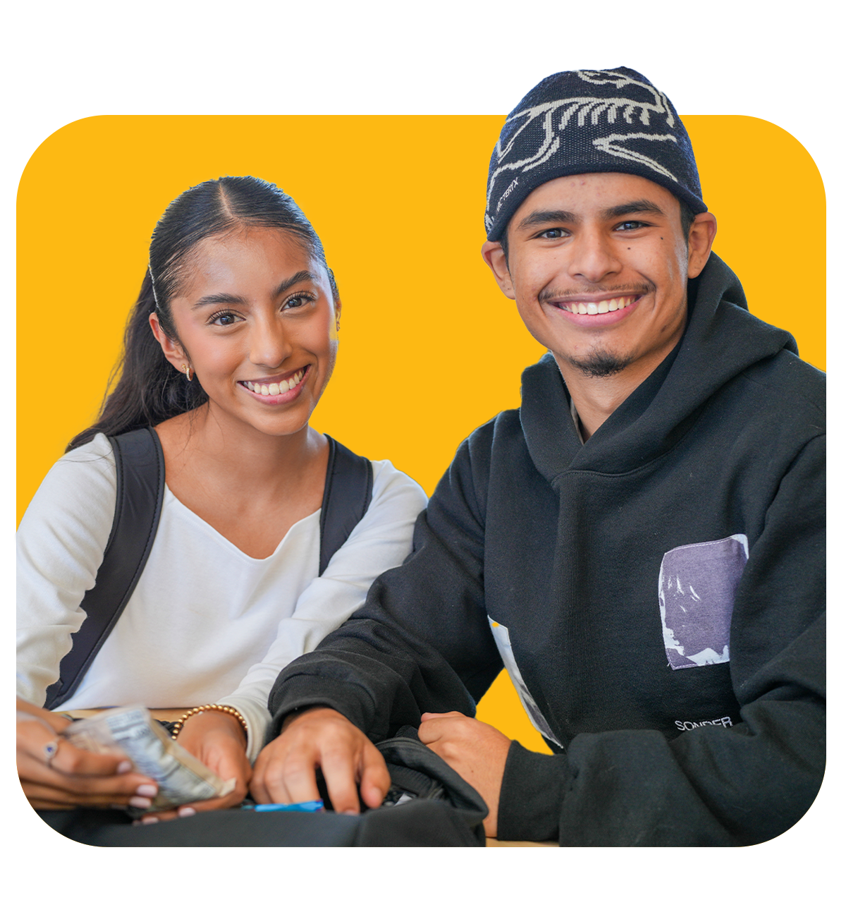 2 hartnell students, a female student on the left sitting down wearing a white sweater and a backpack, smiling at the camera & a male student on the right, wearning a black hoodie and a black beanie, sitting down and smiling at the camera