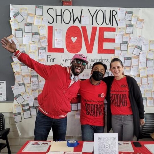 Photo of three faculty members with read shirts, one with a hand raise in excitement, one with a mask, in front of a sign that reads "Show Your Love" with various post-its on it. 