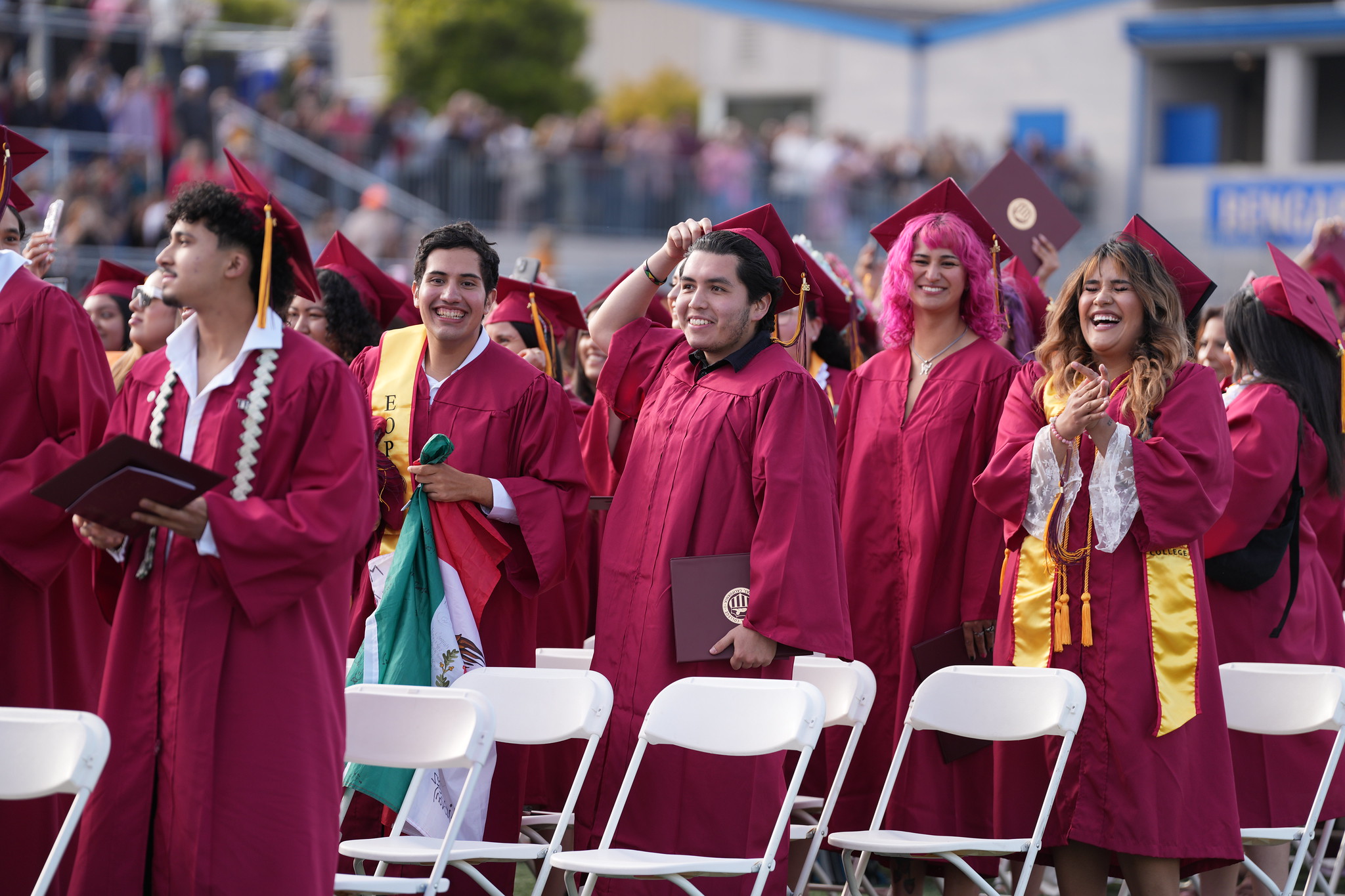 2025 Grads Graduates in maroon caps and gowns stand and celebrate during an outdoor commencement ceremony, smiling and clapping among rows of white chairs.