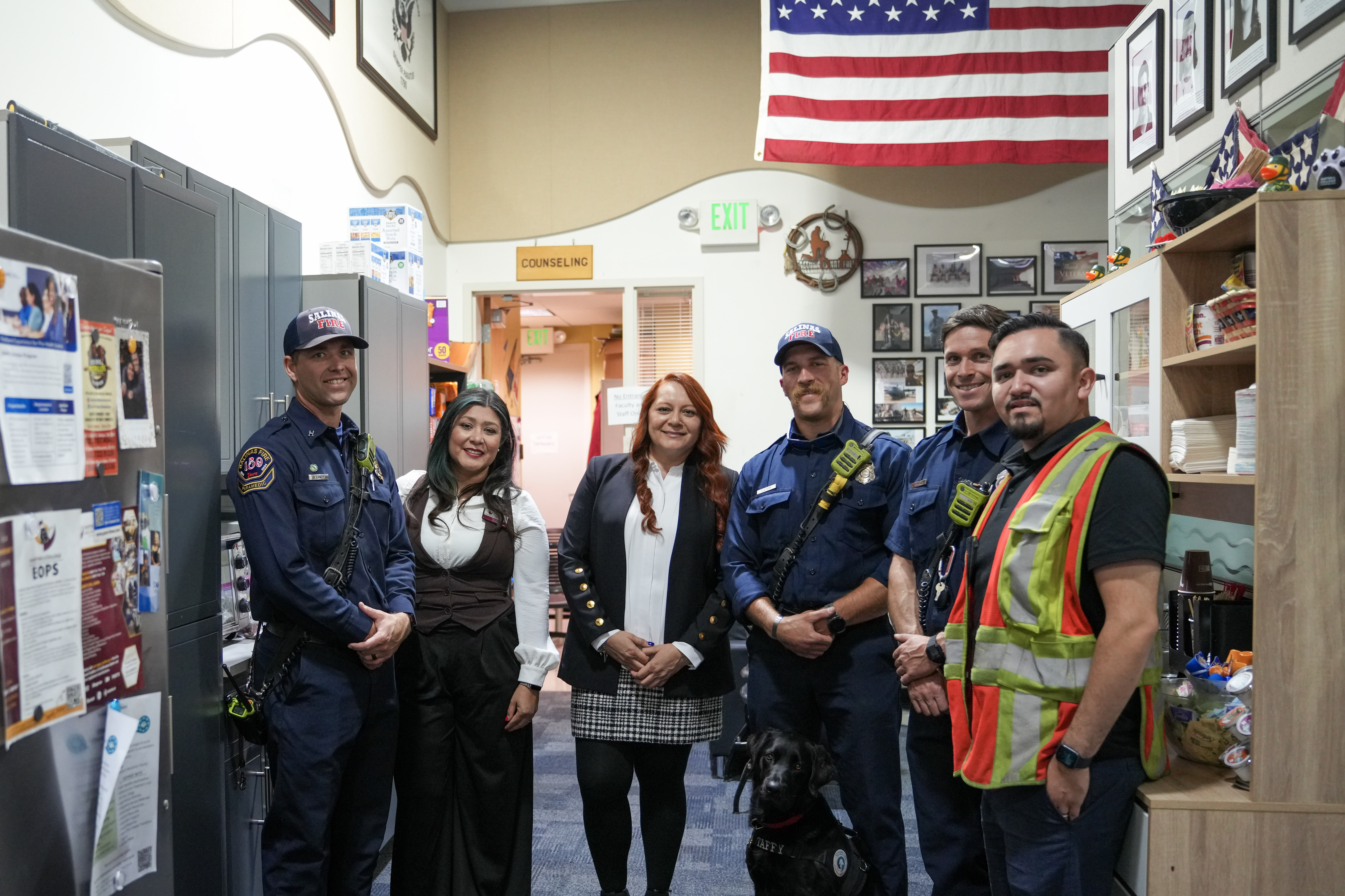 Group of firefighters, community members, and a service dog pose indoors beneath an American flag during a veterans’ outreach event.
