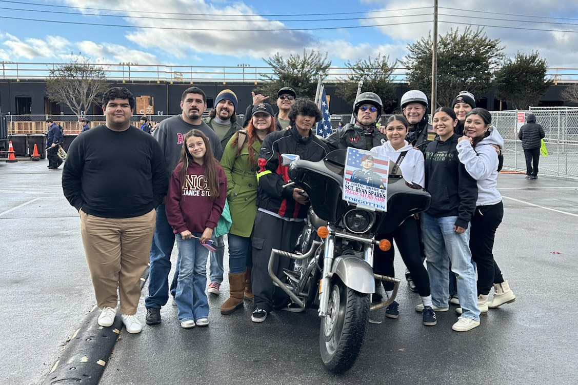 Twelve people posing with motorcyclists and a bike displaying a memorial tribute sign at a public event.