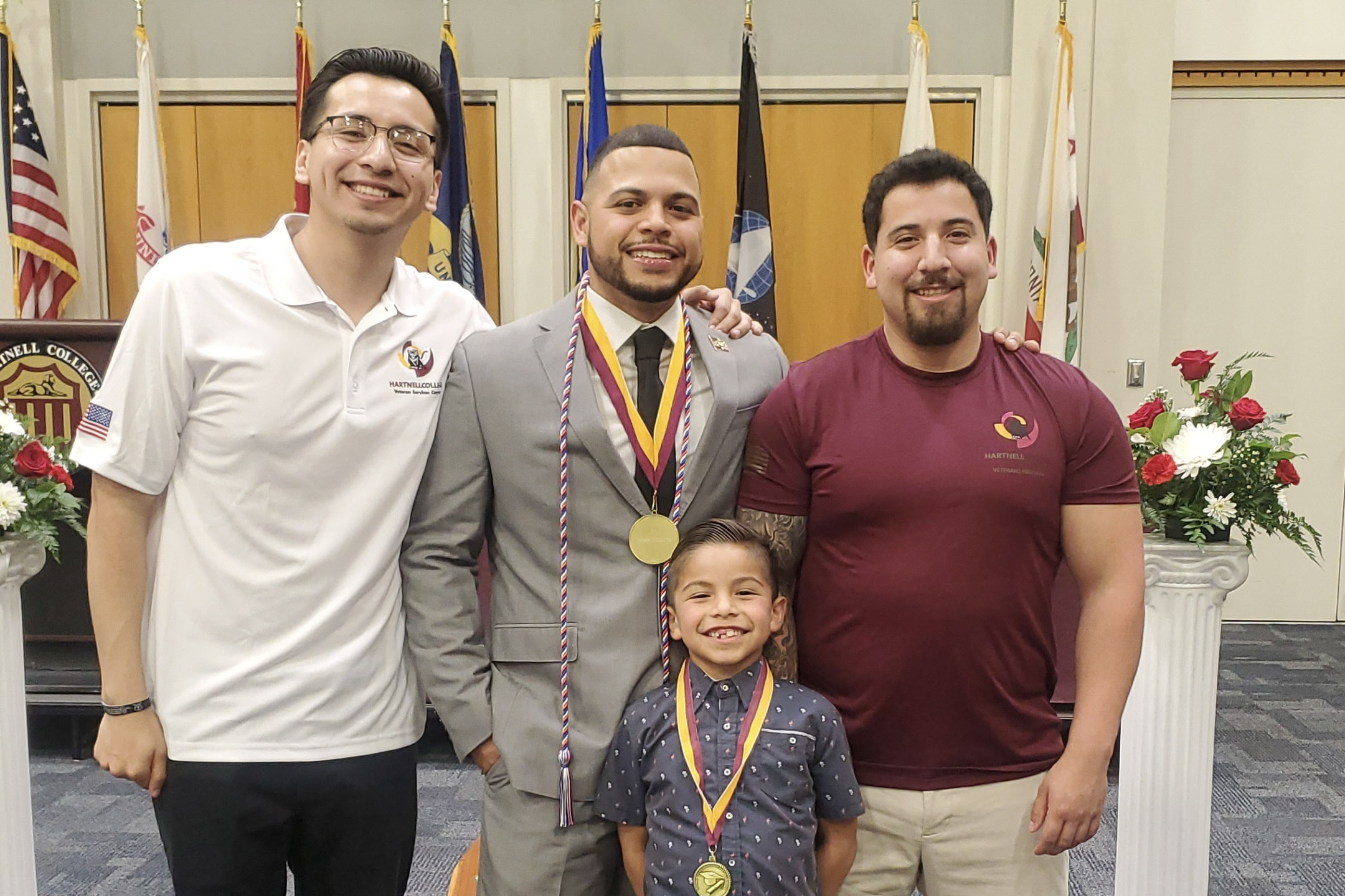 Four people posing indoors at a ceremony, with two wearing medals, one in a suit, one in a white polo, one in a maroon t-shirt, and a smiling young boy in front wearing a medal.