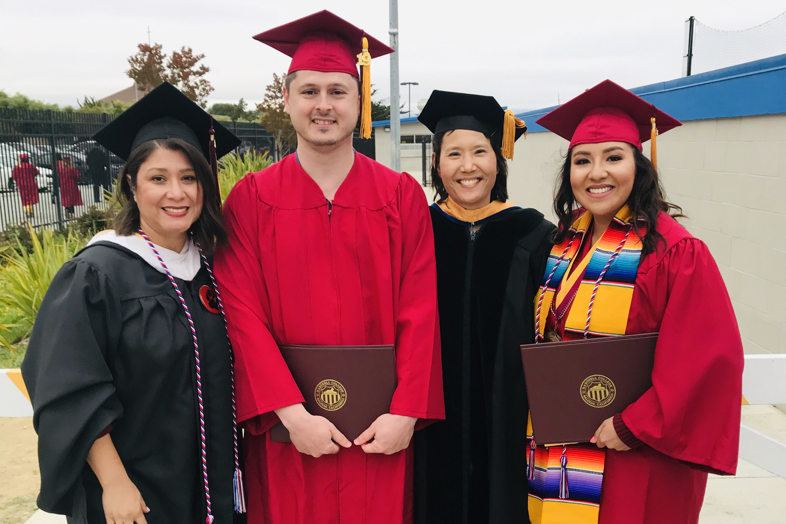 Two graduates in red caps and gowns holding diplomas, standing with two faculty members in black academic regalia, smiling at the camera.