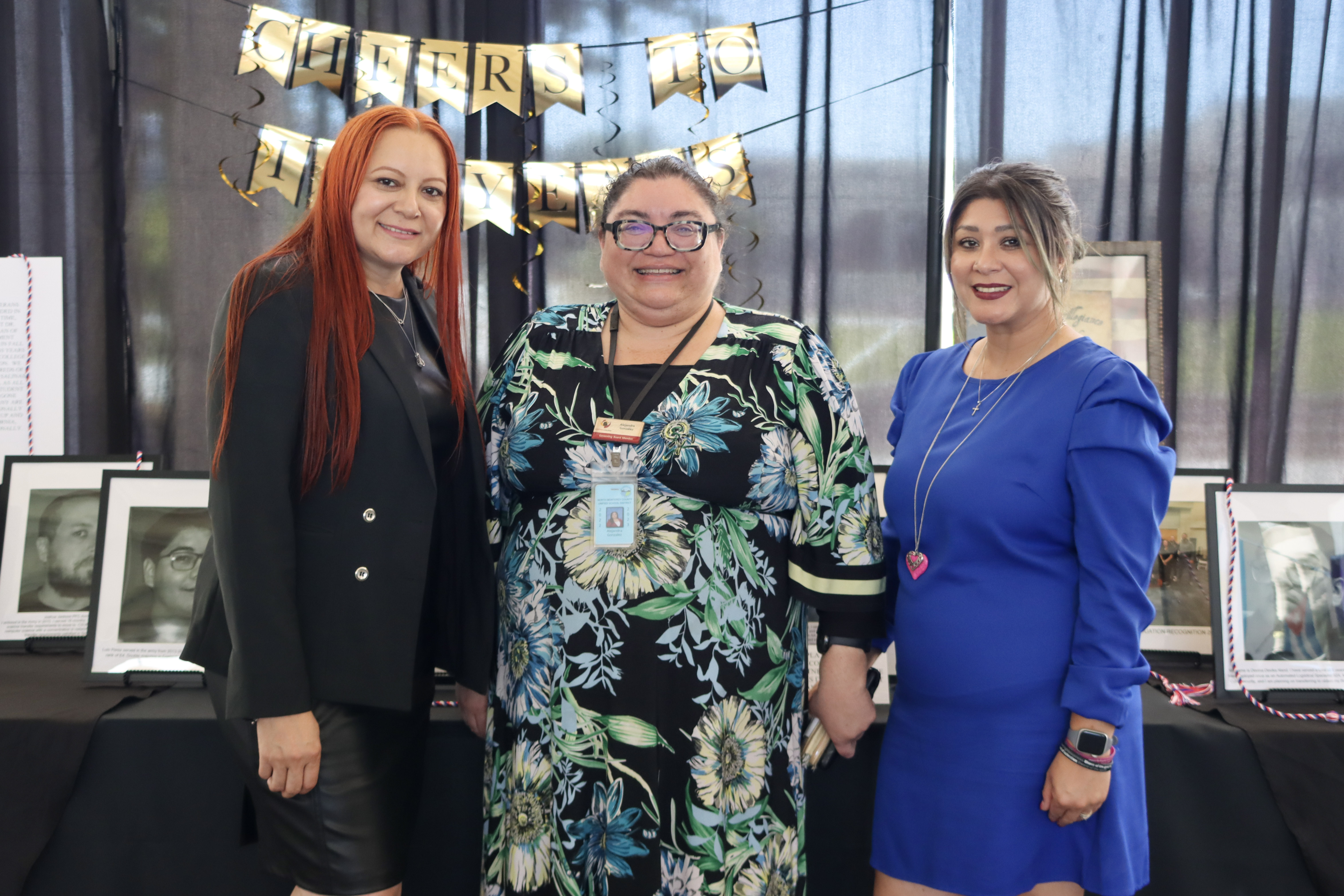 Three women standing together indoors, smiling in front of a black curtain with gold letter banners reading “Cheers” with framed photos on tables behind them.