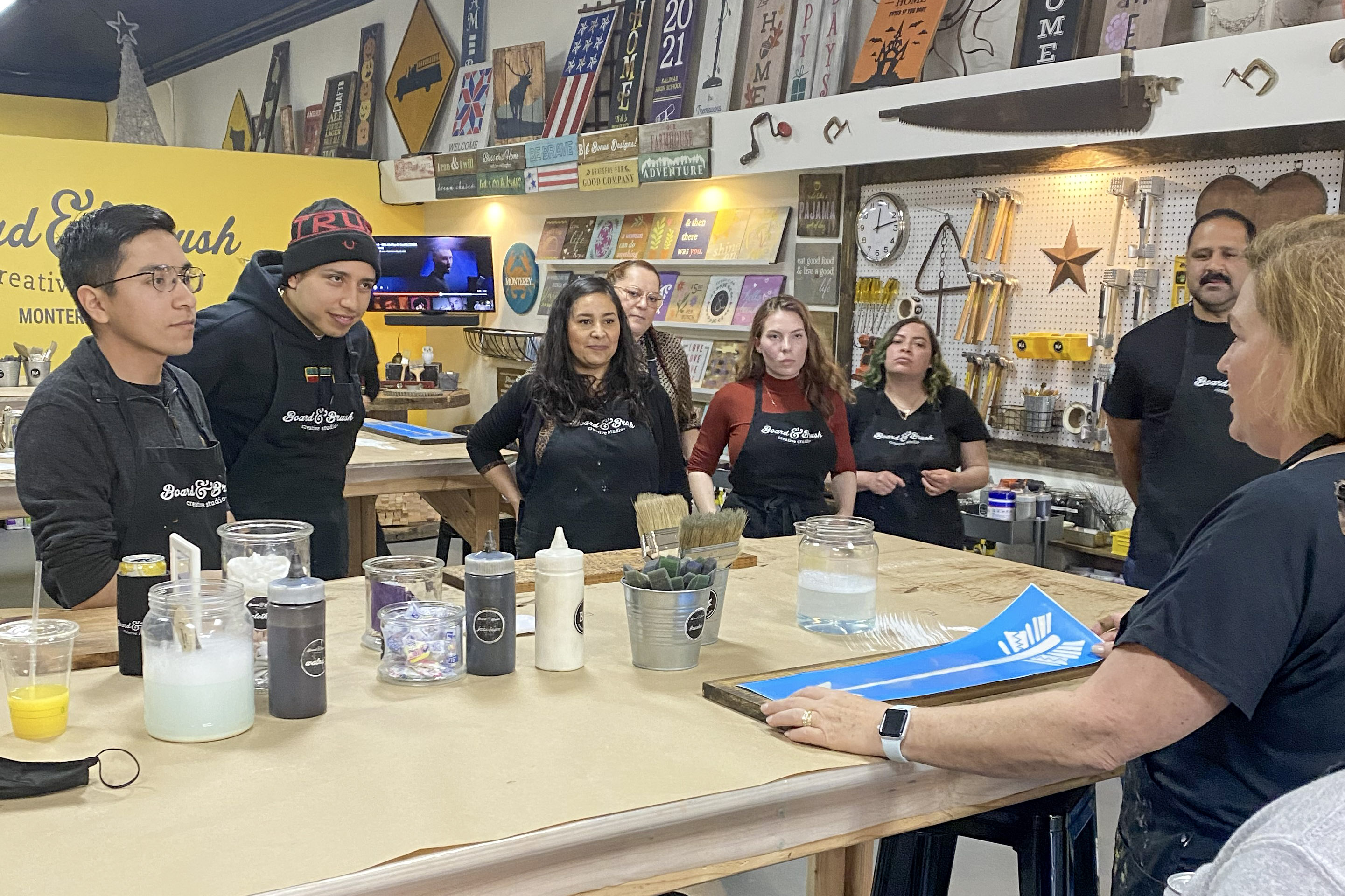 People wearing aprons gathered around a workbench in a creative studio with paint supplies, listening to an instructor.