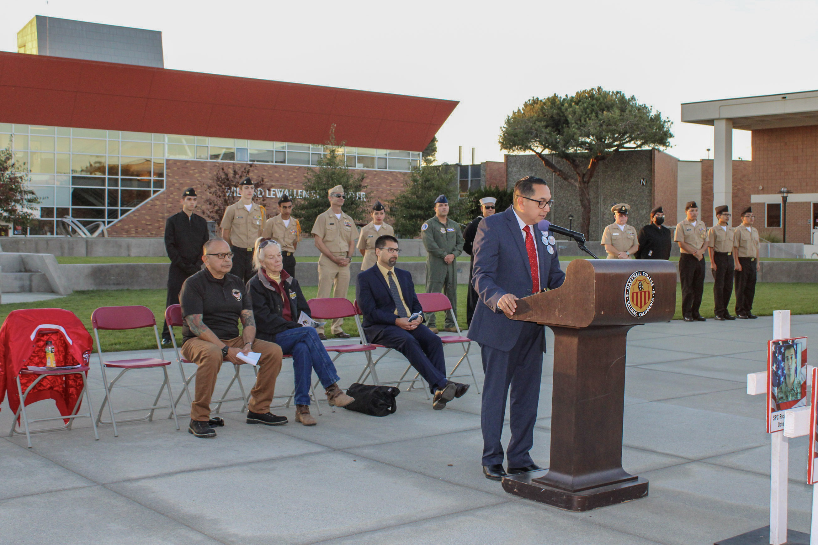 Outdoor ceremony with a man in a suit speaking at a podium, audience members seated, uniformed military personnel standing in formation, and a white cross with a photo in the foreground.
