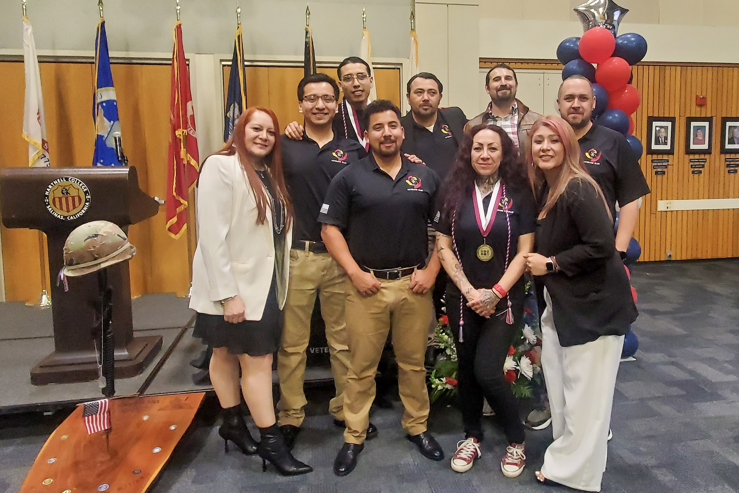 Group of nine people posing indoors in front of military service flags, some wearing medals and cords, standing near a podium and balloon decorations.