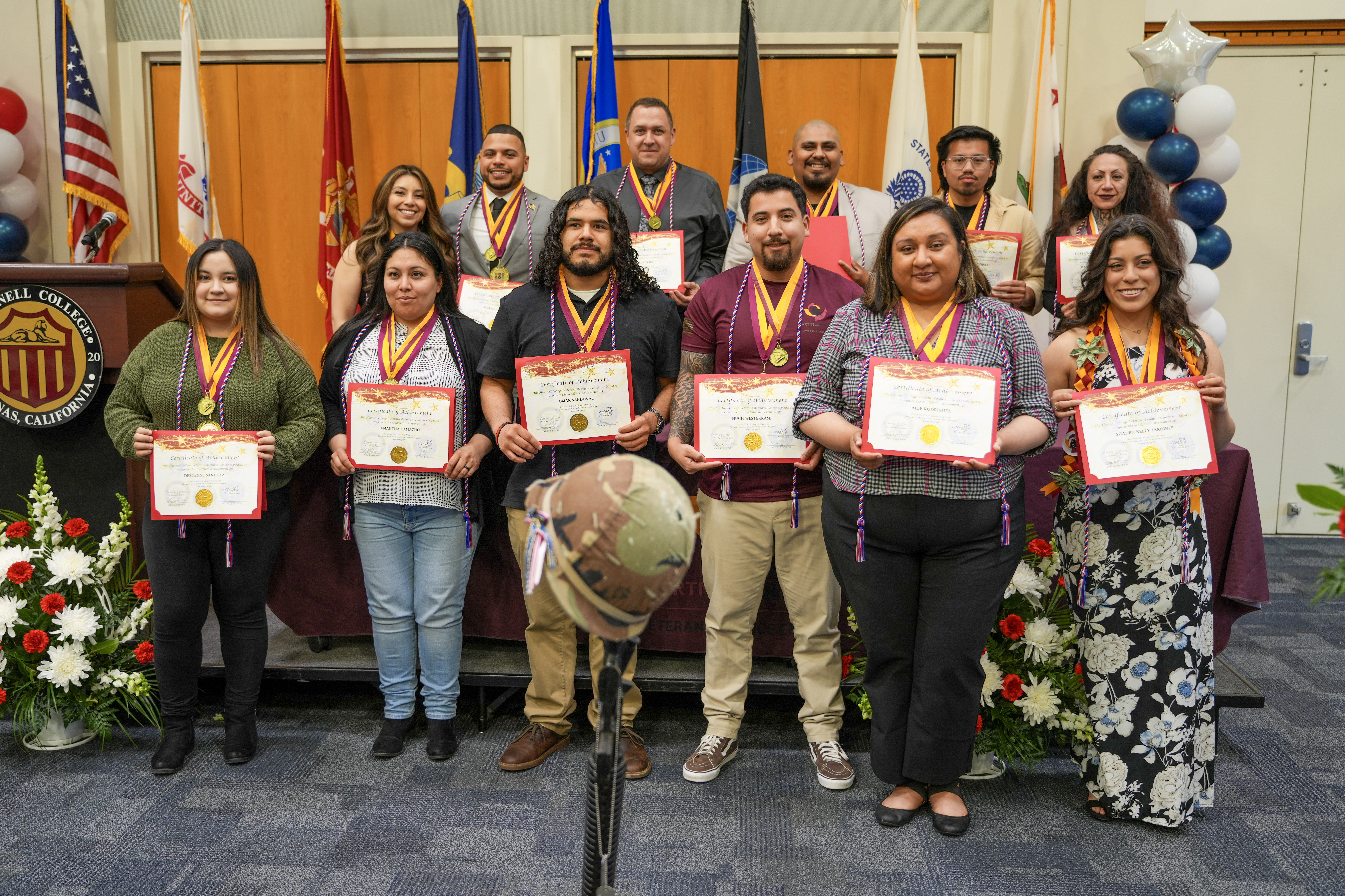 Eleven people standing on stage holding certificates, wearing medals and cords, with military service flags in the background and a helmet on a rifle in the foreground.