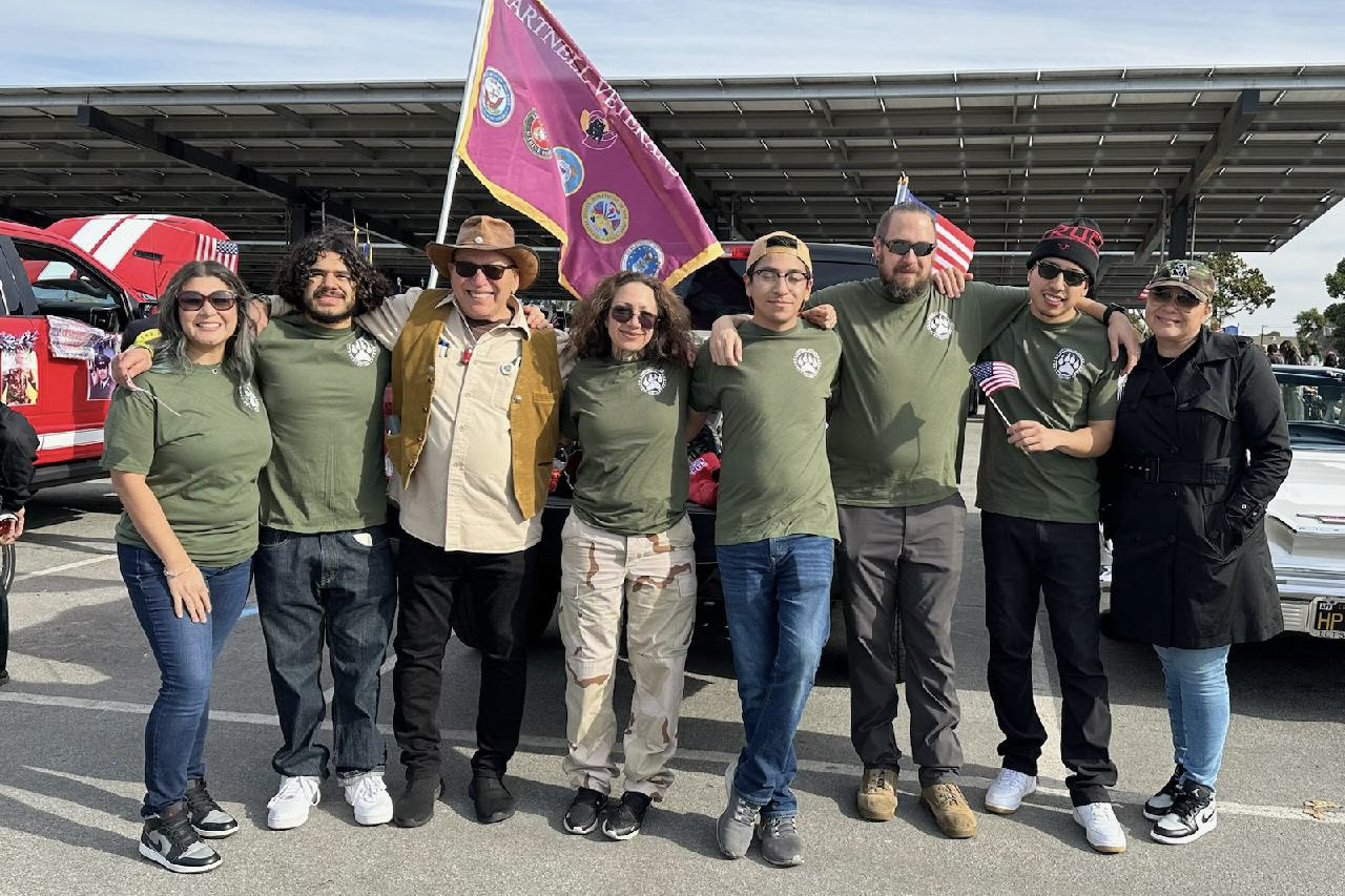Eight smiling people, some in green shirts with a paw print logo, standing together in front of a flag display and vehicles at a public gathering.