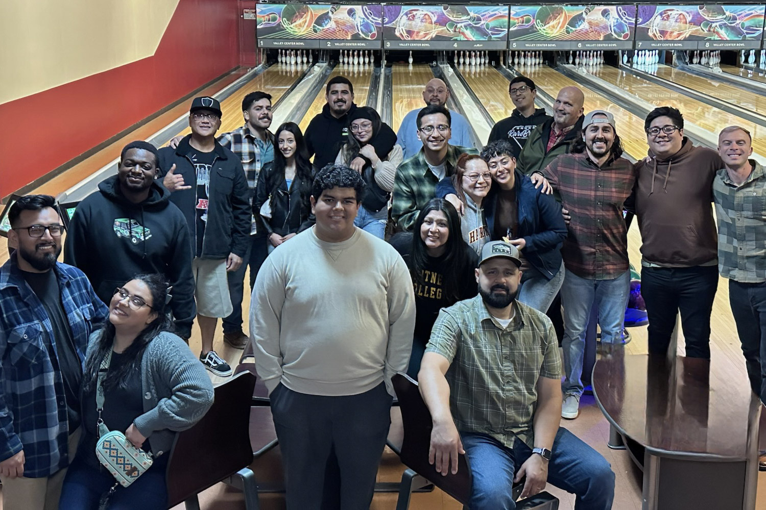 Large group of friends posing at a bowling alley with lanes and pins in the background.