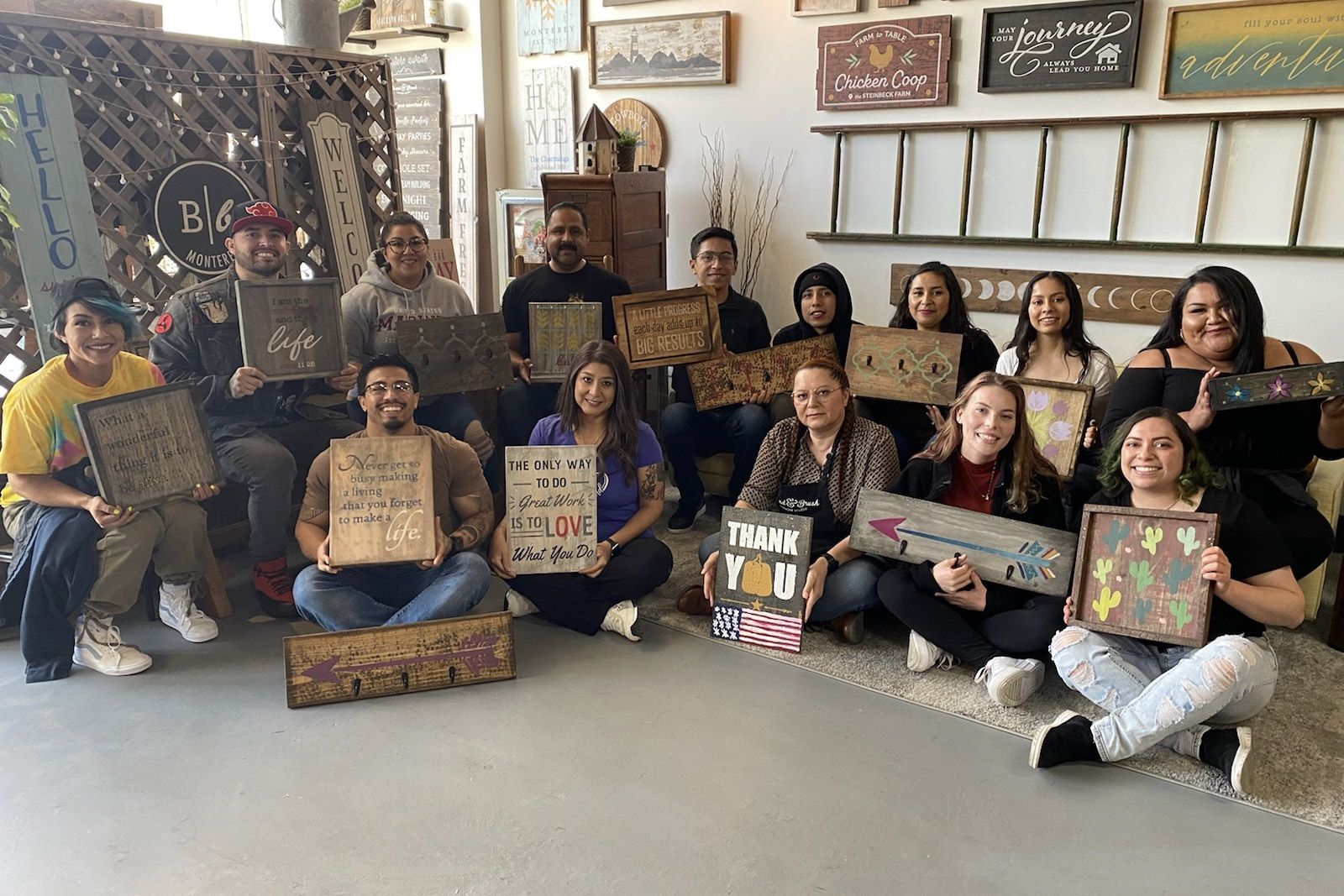 Fifteen people holding handmade wooden signs with inspirational quotes and colorful designs at a craft workshop.